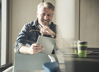 Smiling casual mature businessman sitting at desk using tablet - UUF16730