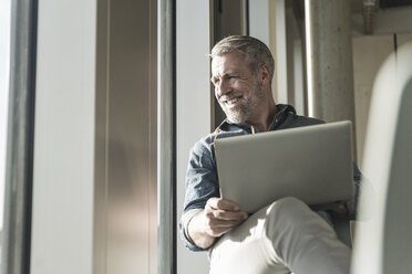 Smiling casual mature businessman with laptop looking out of window - UUF16745