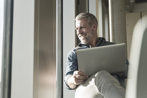 Lächelnder lässiger reifer Geschäftsmann mit Laptop, der aus dem Fenster schaut - UUF16745