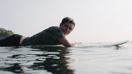 Side view portrait of smiling young man lying on surfboard in sea against sky - CAVF63008