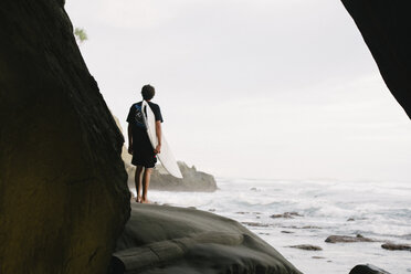 Rückansicht eines Touristen, der auf das Meer blickt, während er ein Surfbrett auf einem Felsen gegen den Himmel hält - CAVF63015