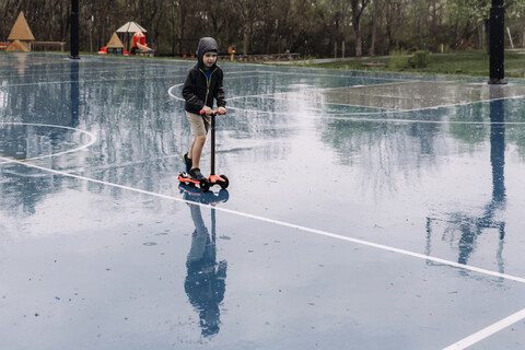 Junge fährt in voller Länge auf einem Roller auf einem nassen Basketballplatz während der Regenzeit, lizenzfreies Stockfoto