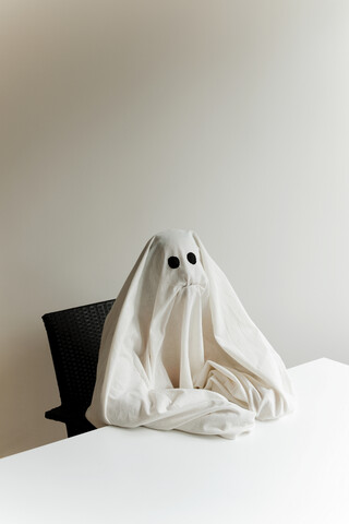 Mann im Gespensterkostüm sitzt zu Hause an einem Tisch an der Wand während Halloween, lizenzfreies Stockfoto