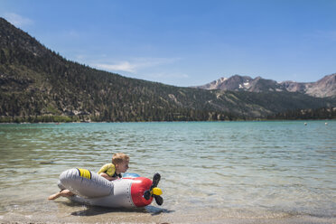 Netter verspielter Junge liegt auf aufblasbarem Flugzeug am See gegen Berge im Sommer - CAVF63120