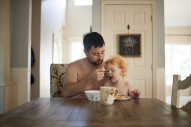 Hemdloser Vater füttert süßen Sohn am Esstisch mit Frühstück - CAVF63130