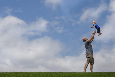 Seitenansicht eines verspielten Vaters, der seinen Sohn in die Luft wirft, während er auf einem grasbewachsenen Feld vor einem bewölkten Himmel steht - CAVF63150