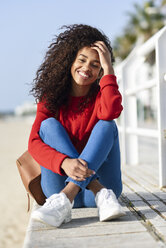 Porträt einer glücklichen jungen Frau, die auf einer Strandpromenade sitzt - JSMF00811