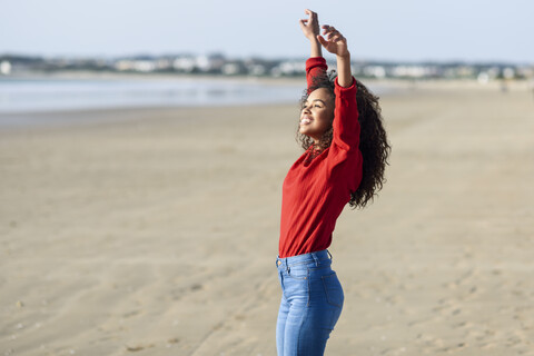 Glückliche junge Frau mit erhobenen Armen am Strand stehend, lizenzfreies Stockfoto