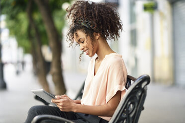 Lächelnde junge Frau, die auf einer Bank sitzt und ein Tablet benutzt - JSMF00851