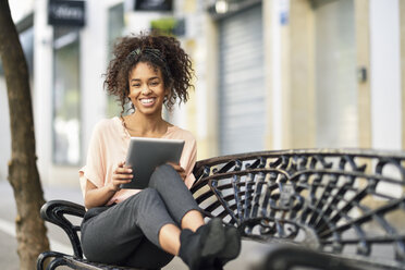 Porträt einer glücklichen jungen Frau, die auf einer Bank sitzt und ein Tablet benutzt - JSMF00854