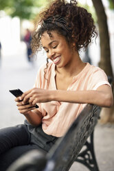 Lächelnde junge Frau sitzt auf einer Bank und benutzt ein Mobiltelefon - JSMF00859