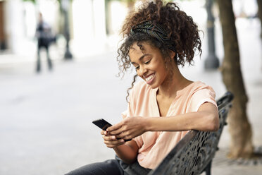 Smiling young woman sitting on a bench using cell phone - JSMF00860