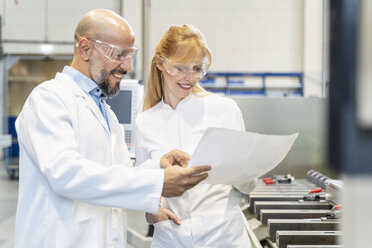 Two happy technicians wearing lab coats and safety glasses looking at plan in factory - DIGF06185