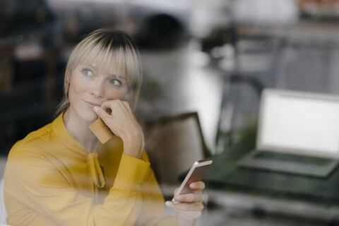 Blonde Geschäftsfrau sitzt am Fenster und bezahlt mit Smartphone und Kreditkarte, lizenzfreies Stockfoto