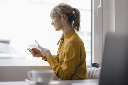 Blonde Geschäftsfrau sitzt am Fenster und bezahlt mit Smartphone und Kreditkarte - JOSF03184