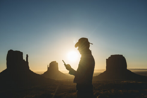 USA, Utah, Monument Valley, Silhouette eines Mannes mit Cowboyhut, der bei Sonnenaufgang auf sein Mobiltelefon schaut - GEMF02898