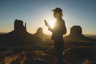 USA, Utah, Monument Valley, Silhouette einer Frau mit Cowboyhut, die bei Sonnenaufgang auf ihr Mobiltelefon schaut - GEMF02899