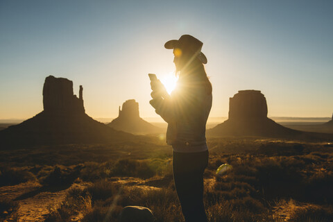 USA, Utah, Monument Valley, Silhouette einer Frau mit Cowboyhut, die bei Sonnenaufgang ein Mobiltelefon benutzt, lizenzfreies Stockfoto