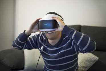 Laughing young man on couch at home using VR glasses - ACPF00487