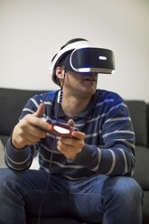 Junger Mann mit VR-Brille sitzt auf der Couch zu Hause und spielt ein Videospiel - ACPF00488