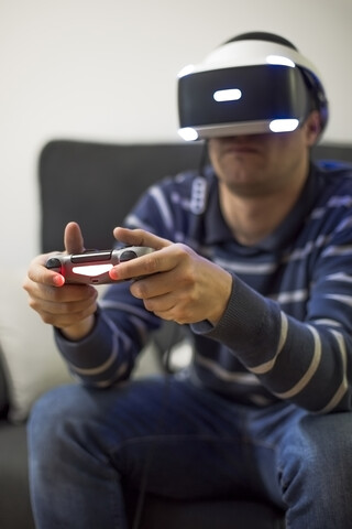Junger Mann mit VR-Brille sitzt auf der Couch zu Hause und spielt ein Videospiel, lizenzfreies Stockfoto