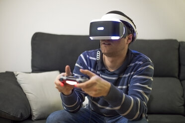 Junger Mann mit VR-Brille sitzt auf der Couch zu Hause und spielt ein Videospiel - ACPF00490