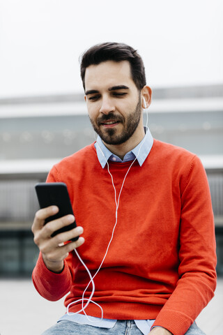 Lässiger Geschäftsmann, der auf einer Bank in der Stadt sitzt und sein Smartphone und Kopfhörer benutzt, lizenzfreies Stockfoto