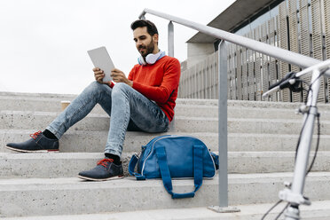 Lässiger Geschäftsmann auf einer Treppe in der Stadt, der sein digitales Tablet und seine Kopfhörer benutzt - JRFF02831