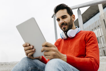 Casual businessman sitting stairs in the city, using his digital tablet and headphones - JRFF02832