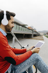 Casual businessman sitting stairs in the city, using his digital tablet and headphones - JRFF02834