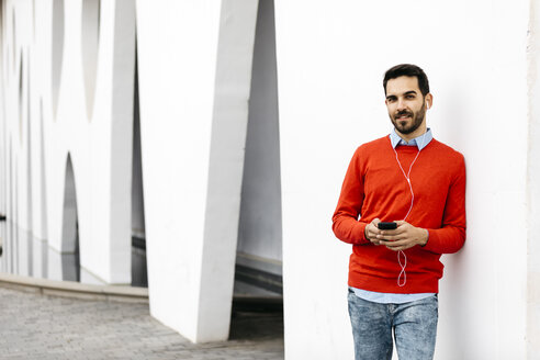 Lässiger Geschäftsmann, der an der Wand lehnt und Kopfhörer und Smartphone benutzt - JRFF02843