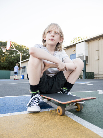 Niedriger Winkel Blick auf nachdenklich Junge mit Skateboard sitzen im Gericht gegen klaren Himmel, lizenzfreies Stockfoto