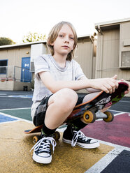 Low angle portrait of boy with skateboard sitting in court against clear sky - CAVF63154