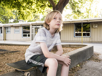 Nachdenklicher Junge mit Skateboard auf Stützmauer im Park sitzend - CAVF63155