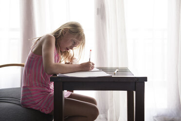 Seitenansicht von niedlichen Mädchen schreiben Hausaufgaben, während auf Stuhl durch Vorhang zu Hause sitzen - CAVF63160