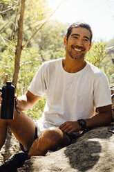 Happy male hiker holding water bottle while sitting on rock against trees in forest - CAVF63164
