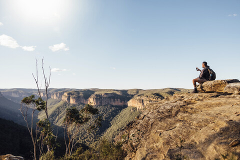 Seitenansicht eines männlichen Wanderers mit Rucksack beim Fotografieren auf einem Berg sitzend gegen den Himmel an einem sonnigen Tag, lizenzfreies Stockfoto