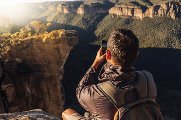 Rückansicht eines männlichen Wanderers mit Rucksack, der auf einem Berg sitzend an einem sonnigen Tag fotografiert - CAVF63170