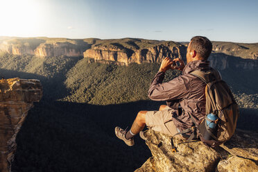 Seitenansicht eines männlichen Wanderers mit Rucksack beim Fotografieren auf einem Berg sitzend gegen den Himmel an einem sonnigen Tag - CAVF63171