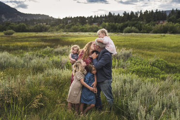Romantische Eltern, die sich küssen, während sie mit ihren Töchtern auf einer Wiese vor dem Himmel im Wald stehen - CAVF63211