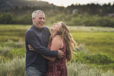 Happy couple with arms around standing on grassy field in forest - CAVF63213