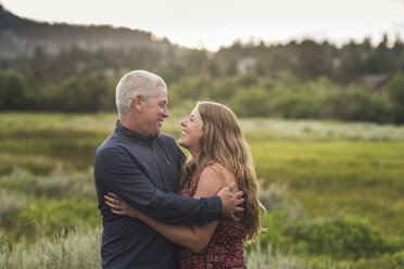 Happy couple with arms around looking at each other while standing on grassy field in forest - CAVF63214