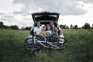 Glückliche Familie mit Fahrrädern, die auf dem Kofferraum eines Autos sitzen, inmitten eines grasbewachsenen Feldes gegen den bewölkten Himmel - CAVF63229