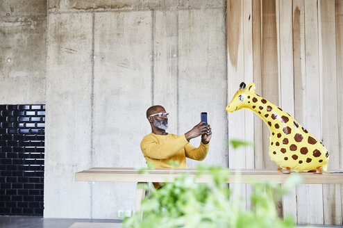 Älterer Geschäftsmann sitzt am Schreibtisch im Büro mit Handy und Giraffenfigur - FMKF05454