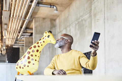 Älterer Geschäftsmann sitzt am Schreibtisch im Büro mit Handy und küsst Giraffe Figur, lizenzfreies Stockfoto