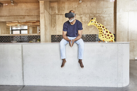 Mature businessman in modern office sitting next to giraffe figurine wearing VR glasses stock photo