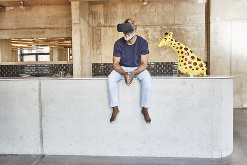 Mature businessman in modern office sitting next to giraffe figurine wearing VR glasses - FMKF05472