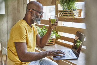 Älterer Geschäftsmann mit Smoothie und Laptop am Fenster im Büro - FMKF05476