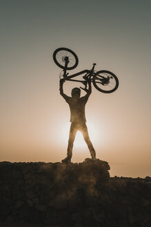 Spanien, Lanzarote, Mountainbiker auf einem Ausflug bei Sonnenuntergang, der sein Fahrrad hochhebt - AHSF00103