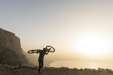 Spain, Lanzarote, mountainbiker on a trip at the coast at sunset carrying his bike - AHSF00108